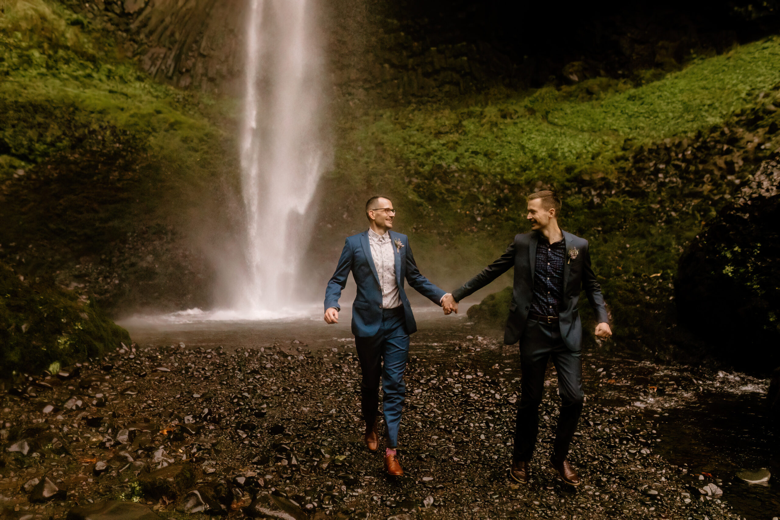 Two grooms hold hands while running in front of Latourell Falls after their elopement ceremony.